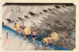 Driving Rain at Shōno by Hiroshige: travellers in straw capes rushing along a slope in slanting rain, bamboo bending in grey