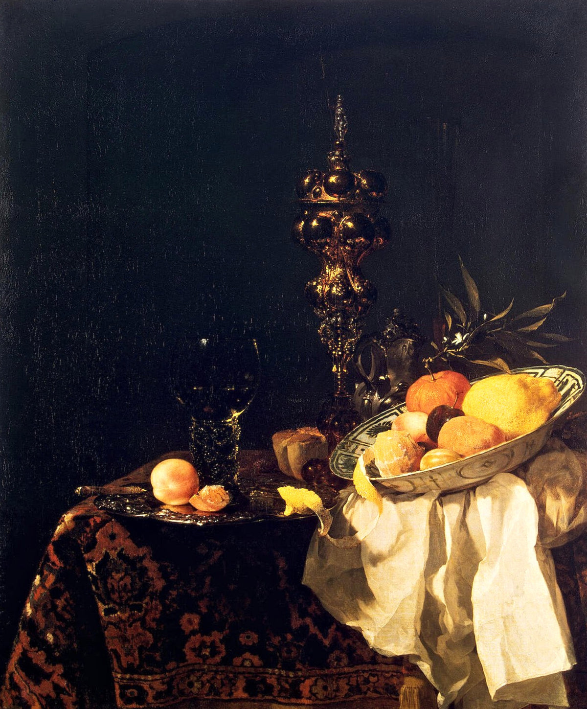 Dessert by Willem Kalf: a gilt cup, a wine glass and a silver dish of oranges and lemons on a Turkish carpet, a white napkin